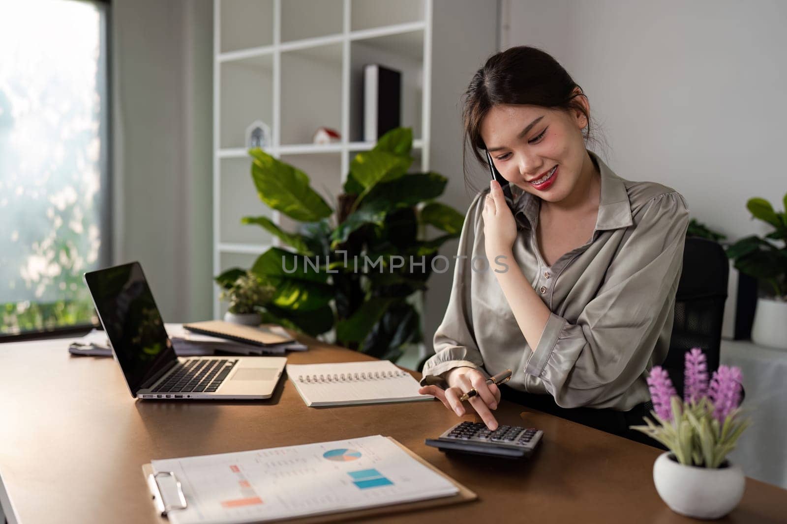 Young woman working from home office, using a calculator and smiling, surrounded by modern office supplies and indoor plants in a bright, minimalist workspace.