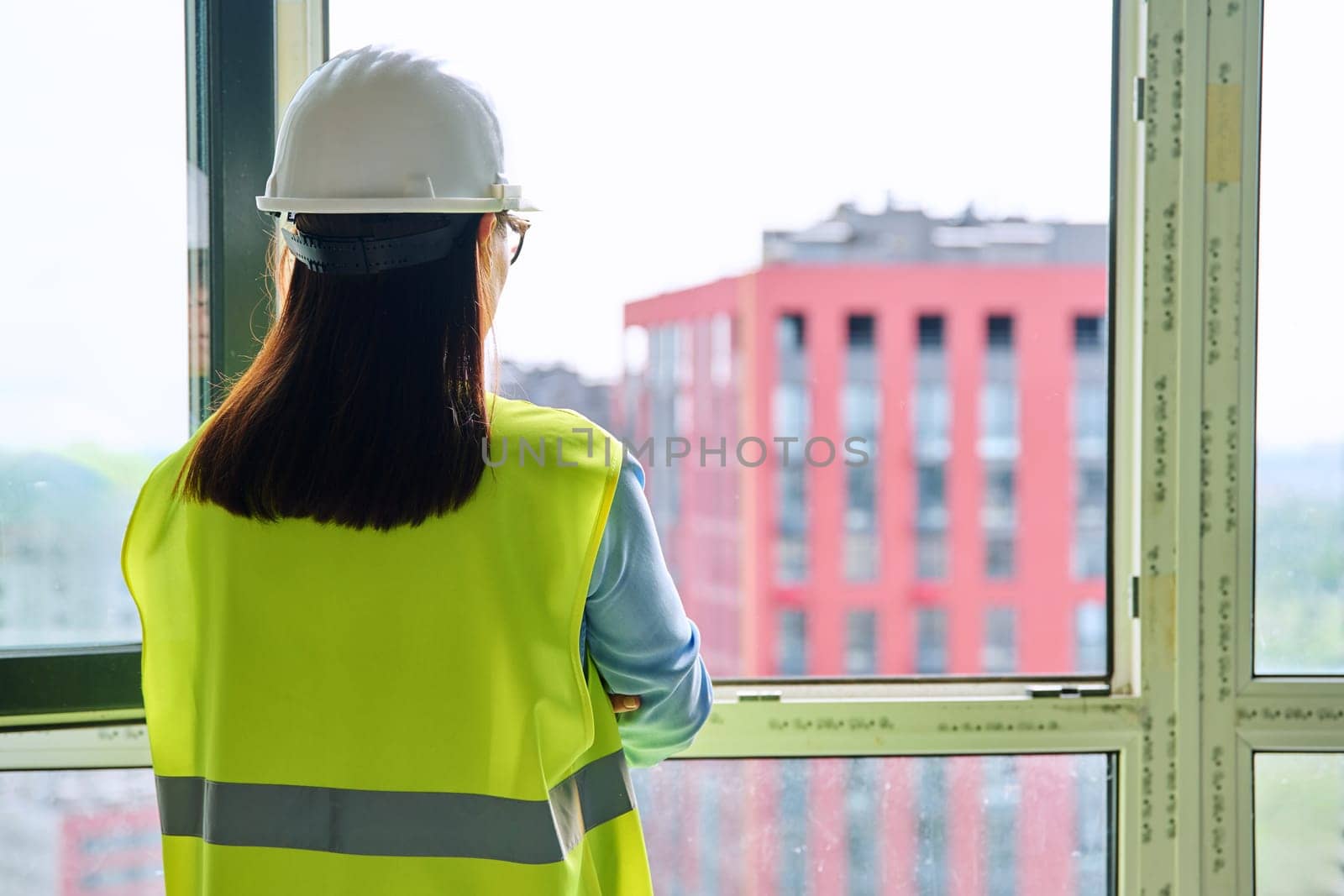 Back view of female industrial worker in protective helmet vest looking at new buildings through window. Construction engineering, real estate sales buying office apartment, work technical professions