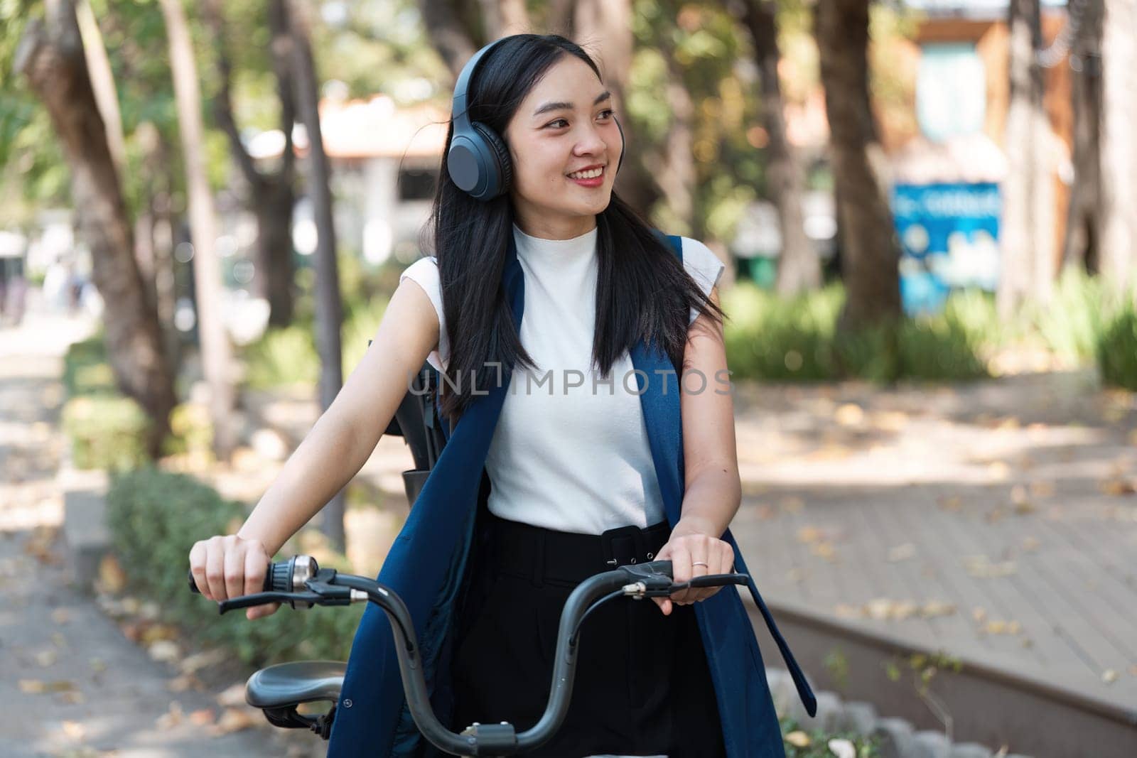 Young woman riding a bicycle in an urban park, embracing an eco-friendly lifestyle. She is smiling, wearing casual clothing and headphones, surrounded by nature.