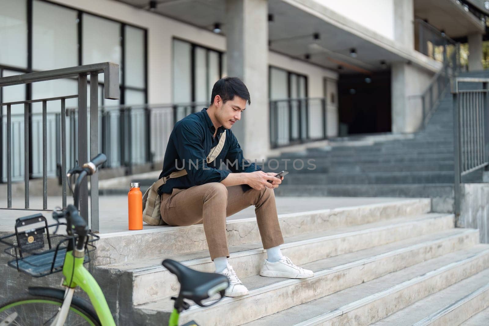 A young professional using a smartphone while sitting on steps next to a green bicycle, embodying a business eco lifestyle in an urban environment.