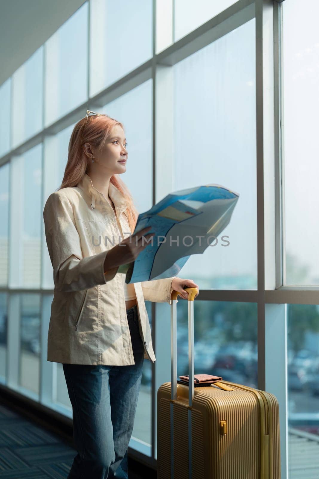 A woman stands in an airport terminal holding a map and suitcase, ready for her travel adventure. She looks out the window, planning her journey.