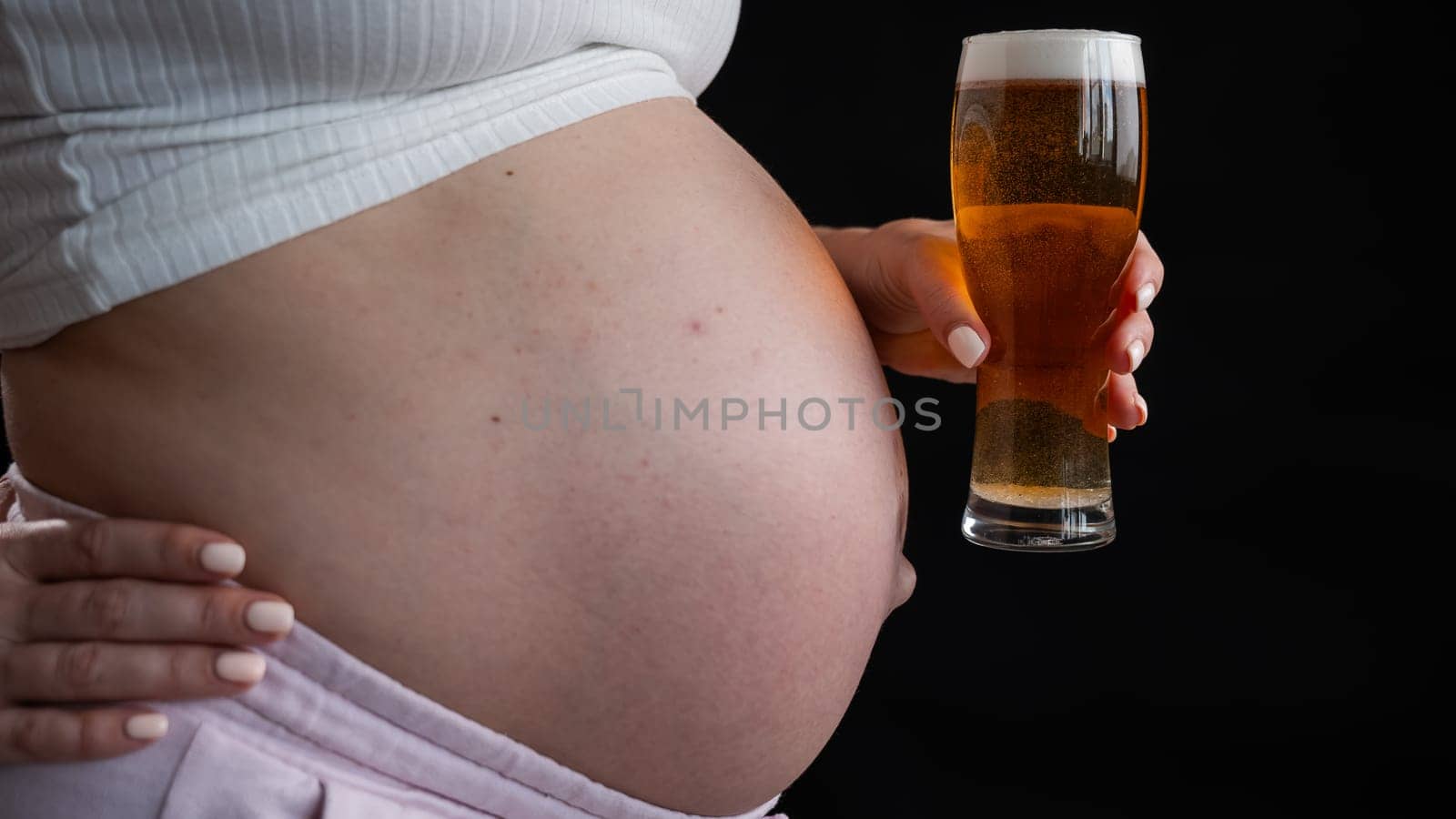 Close-up of the belly of a pregnant woman holding a glass of beer on a black background. Skin rash. by mrwed54