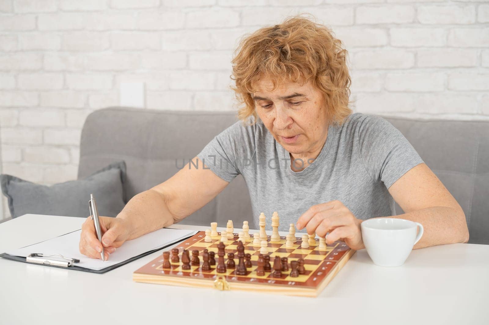 Elderly Caucasian woman writes down moves in a notebook while playing chess. by mrwed54