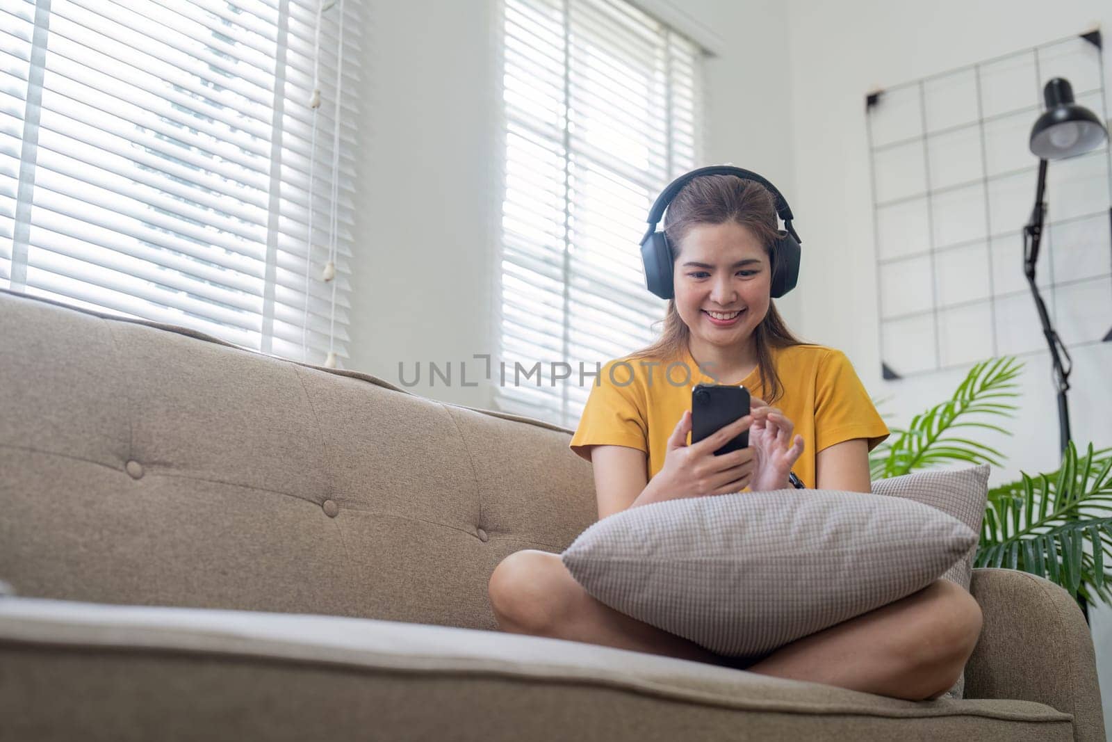 A young woman wearing headphones, sitting on a sofa, and enjoying music on her smartphone in a bright and modern living room.