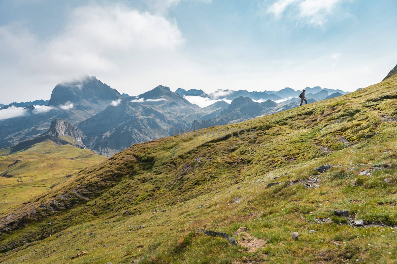 A person is walking up a grassy hill in the mountains under a cloudy sky, surrounded by lush greenery with a vast landscape spreading in the horizon. High quality photo