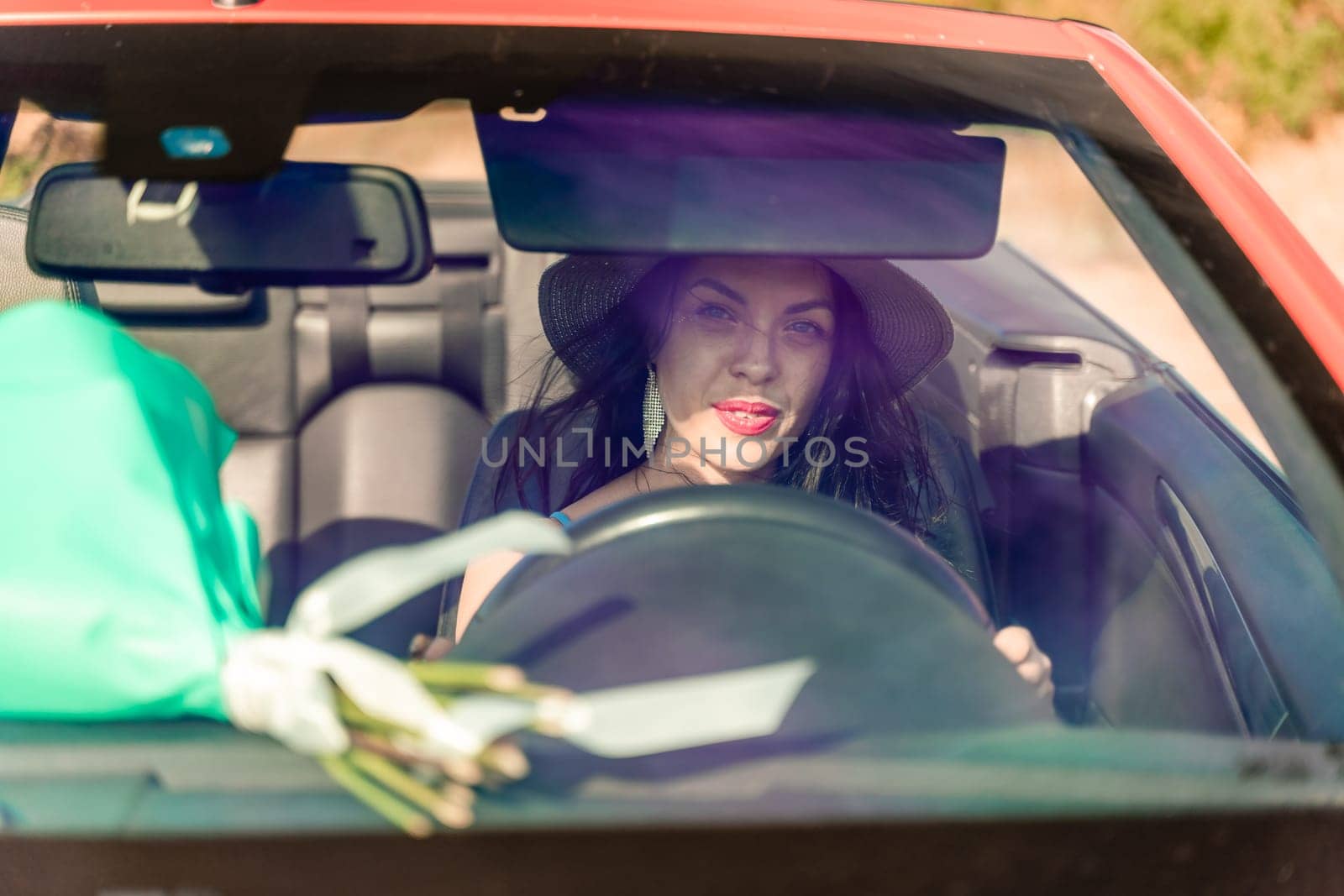 A woman is driving a car with a bouquet of flowers in the back seat. She is wearing a hat and has her eyes closed