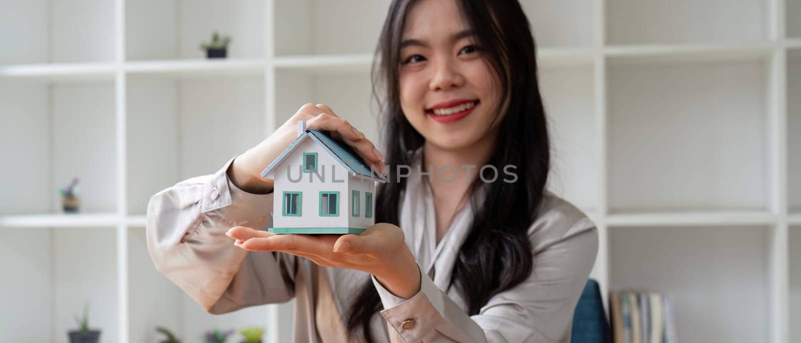 Smiling Woman Holding Miniature House Model in Hands, Real Estate Agent Promoting Property Sale, Modern Home Concept, Professional Realtor Offering New Home, Real Estate Investment Opportunity by nateemee