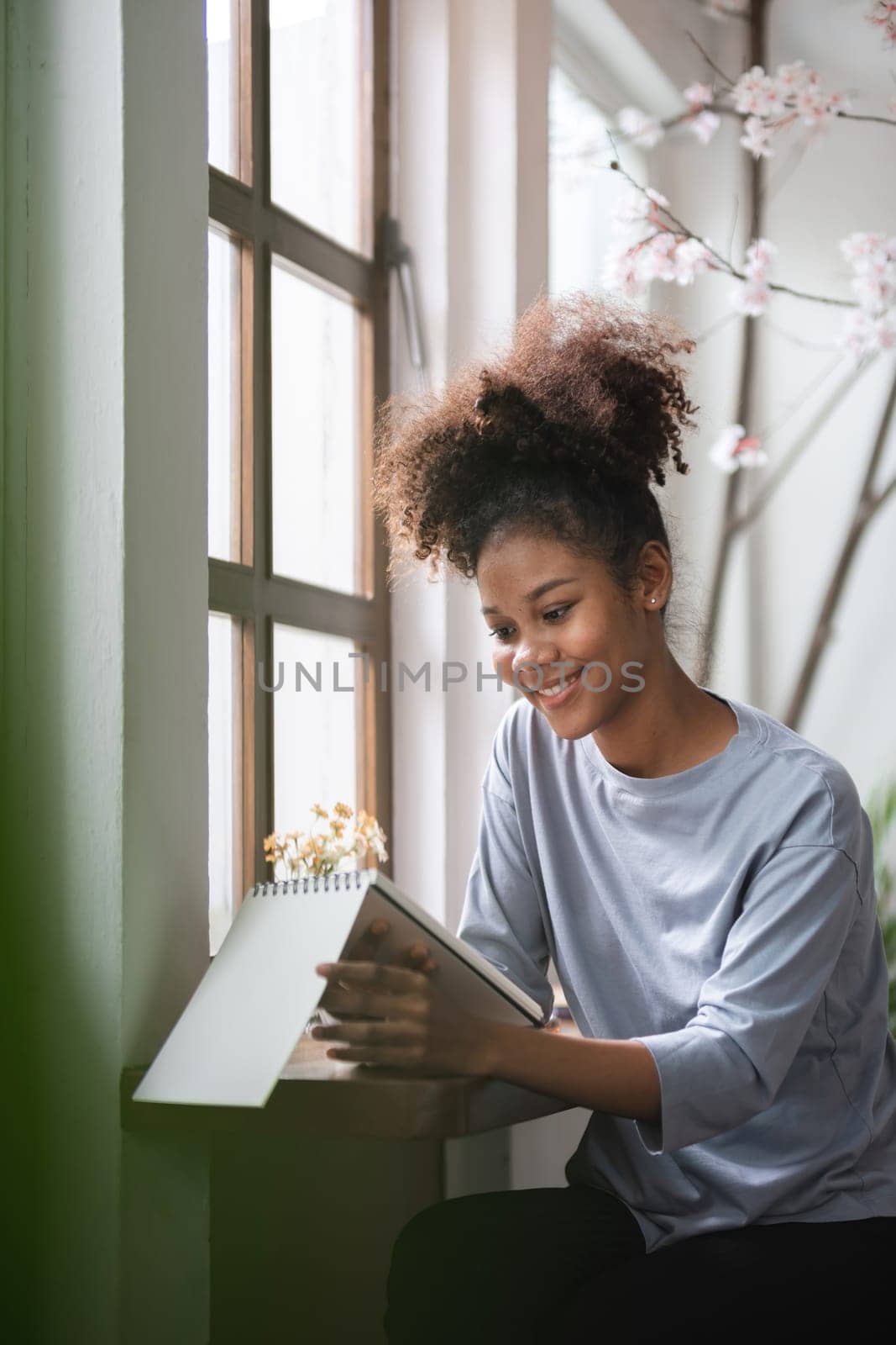 Young Woman Sitting in Modern Living Room Taking Notes by the Window, Smiling and Engaged in Creative Work, Natural Light, Cozy Home Environment, Casual Clothing, Productive and Relaxed Atmosphere by wichayada