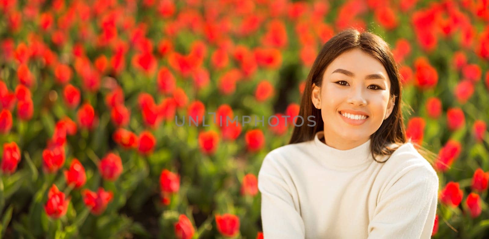 Asian woman is standing in front of a vast field filled with vibrant red flowers. She is looking out at the flowers, the sun shining down on her as she takes in the natural beauty of the scene.