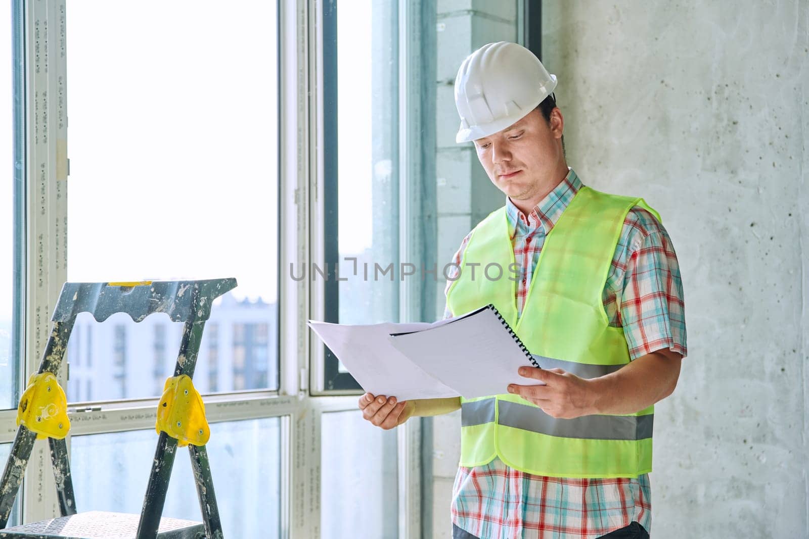 Portrait of young male industrial worker in safety vest helmet with working engineering architectural documents on construction. Construction repair renovation service development work concept