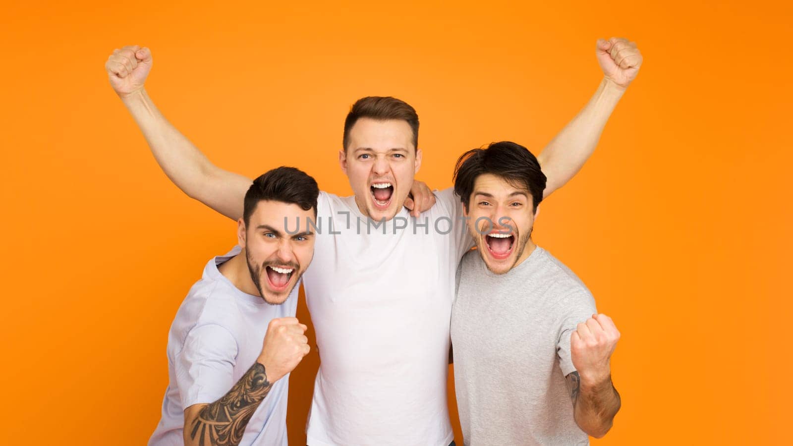 Three young men are exuberantly celebrating, arms raised in triumph, against a vibrant orange background