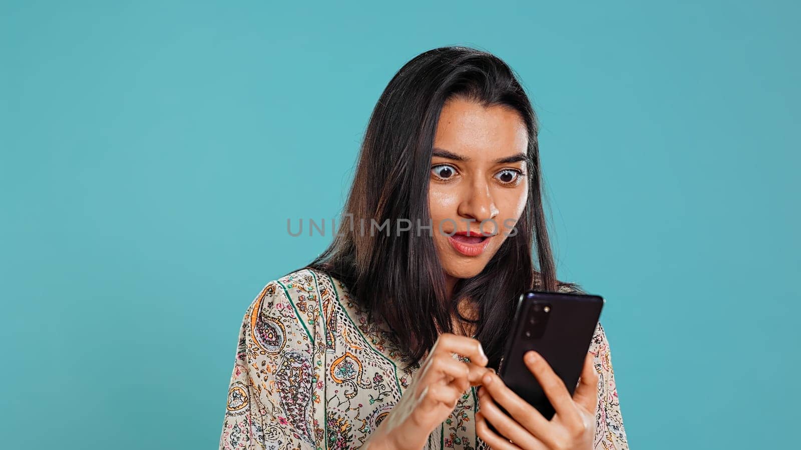 Woman happily scrolling on phone touchscreen to check social media feed, amazed by photo. Indian person browsing internet websites, astonished by content, studio background, camera B