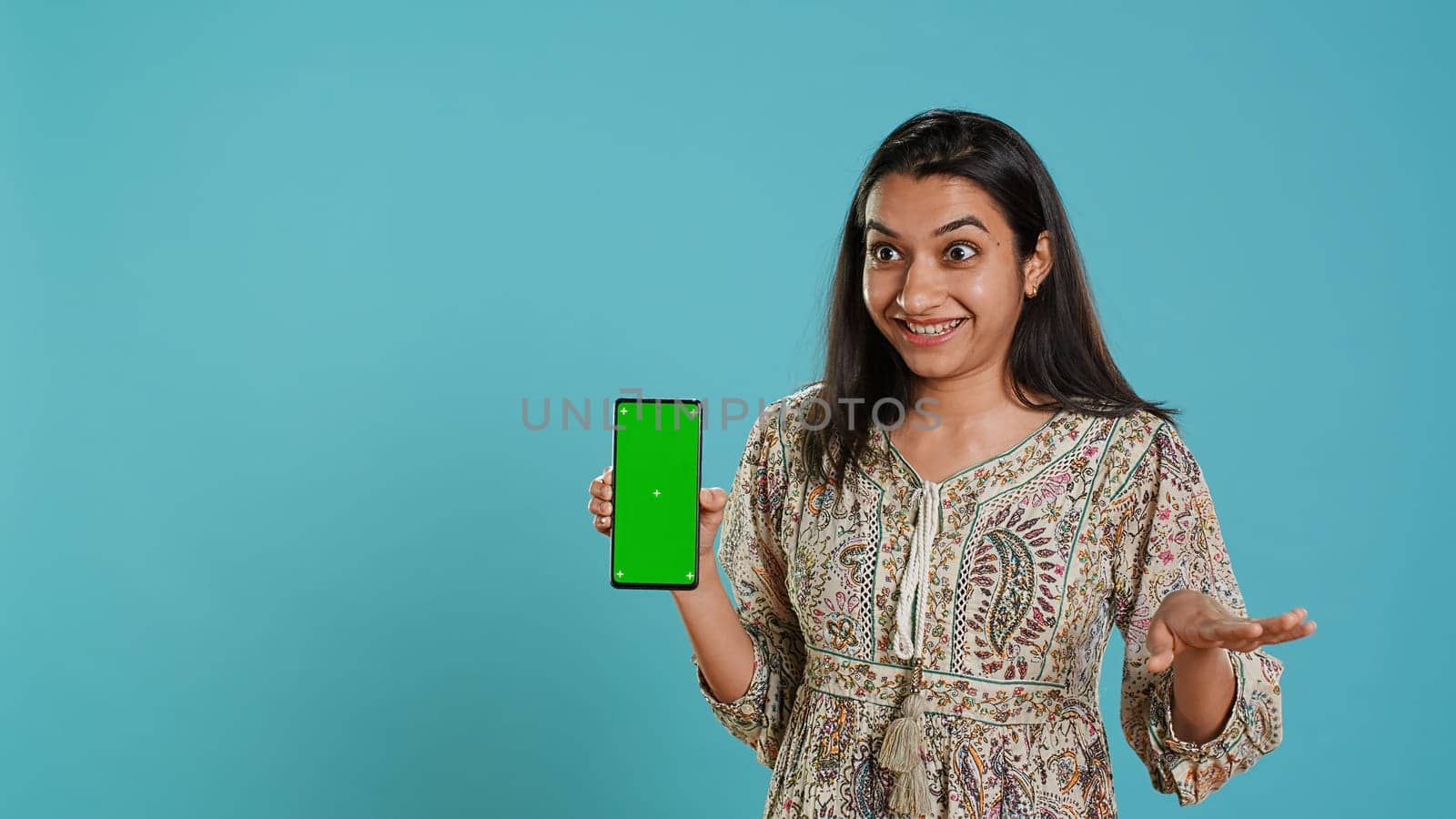 Tech content creator doing influencer marketing using green screen smartphone, isolated over studio background. Indian woman holding empty copy space mockup phone, doing review, camera A