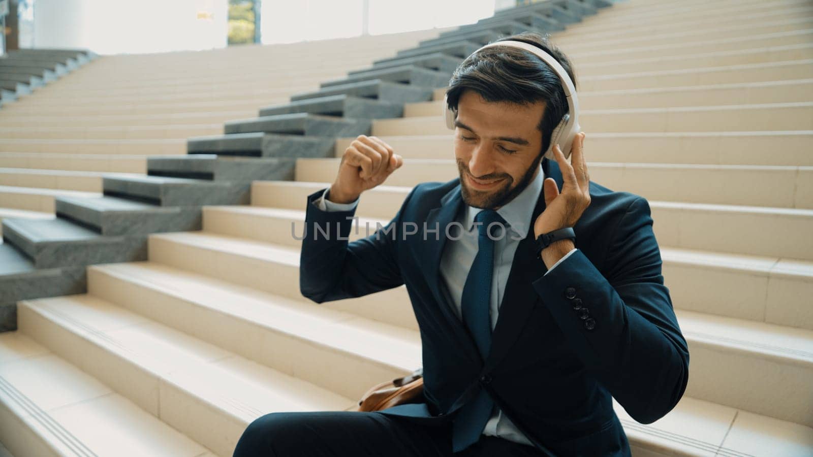 Project manager listening and enjoy music from headphone while sitting at stairs. Professional businessman wearing suit outfit while dancing and moving to music. Happy man listen funny song. Exultant.