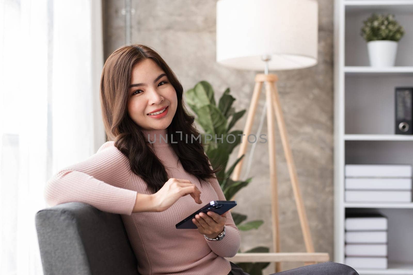 A young woman sits comfortably on a sofa in a modern living room, smiling while using her smartphone. The room features contemporary design elements, natural light, and an indoor plant.