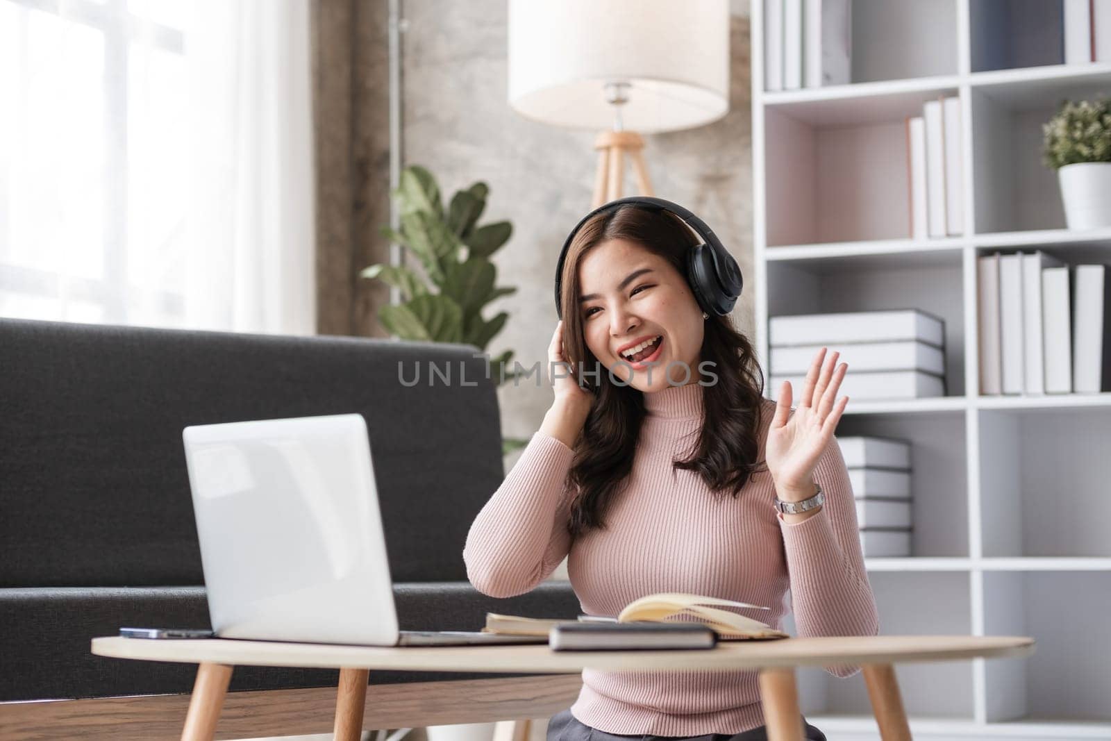 Young woman working in a modern living room, using a laptop and headphones, smiling and waving, surrounded by books and cozy decor.