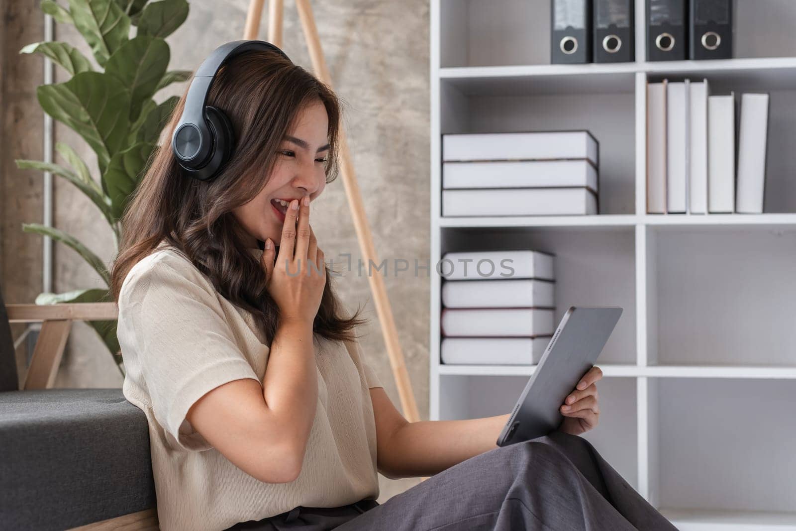 A young woman wearing headphones, using a tablet, and sitting in a modern living room with stylish decor and shelves.