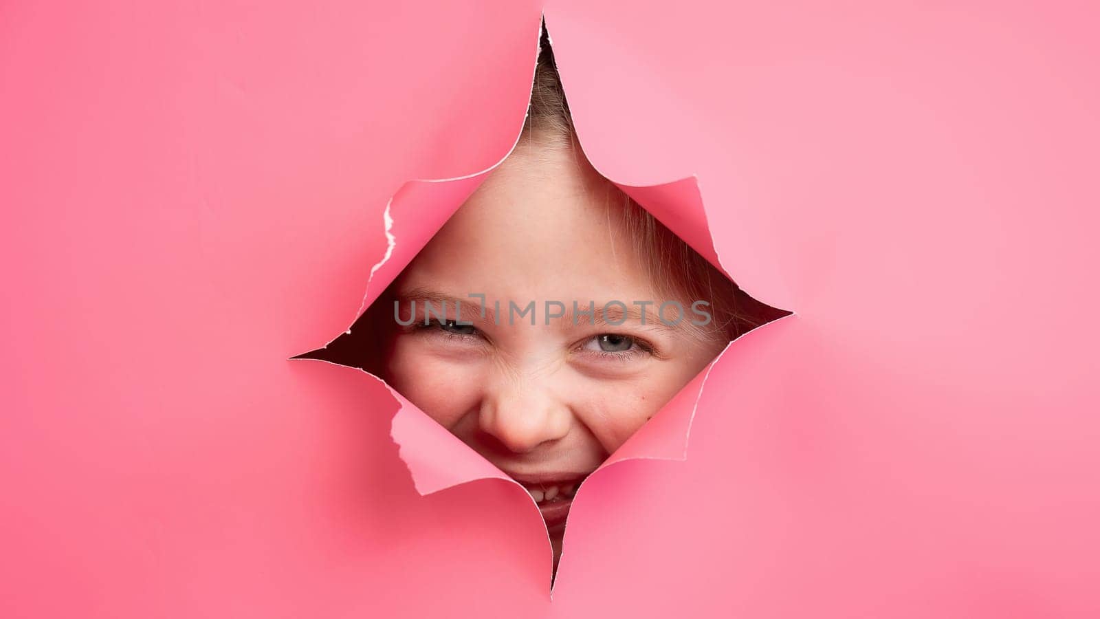 Cute Caucasian girl peeks out of a hole in a paper pink background