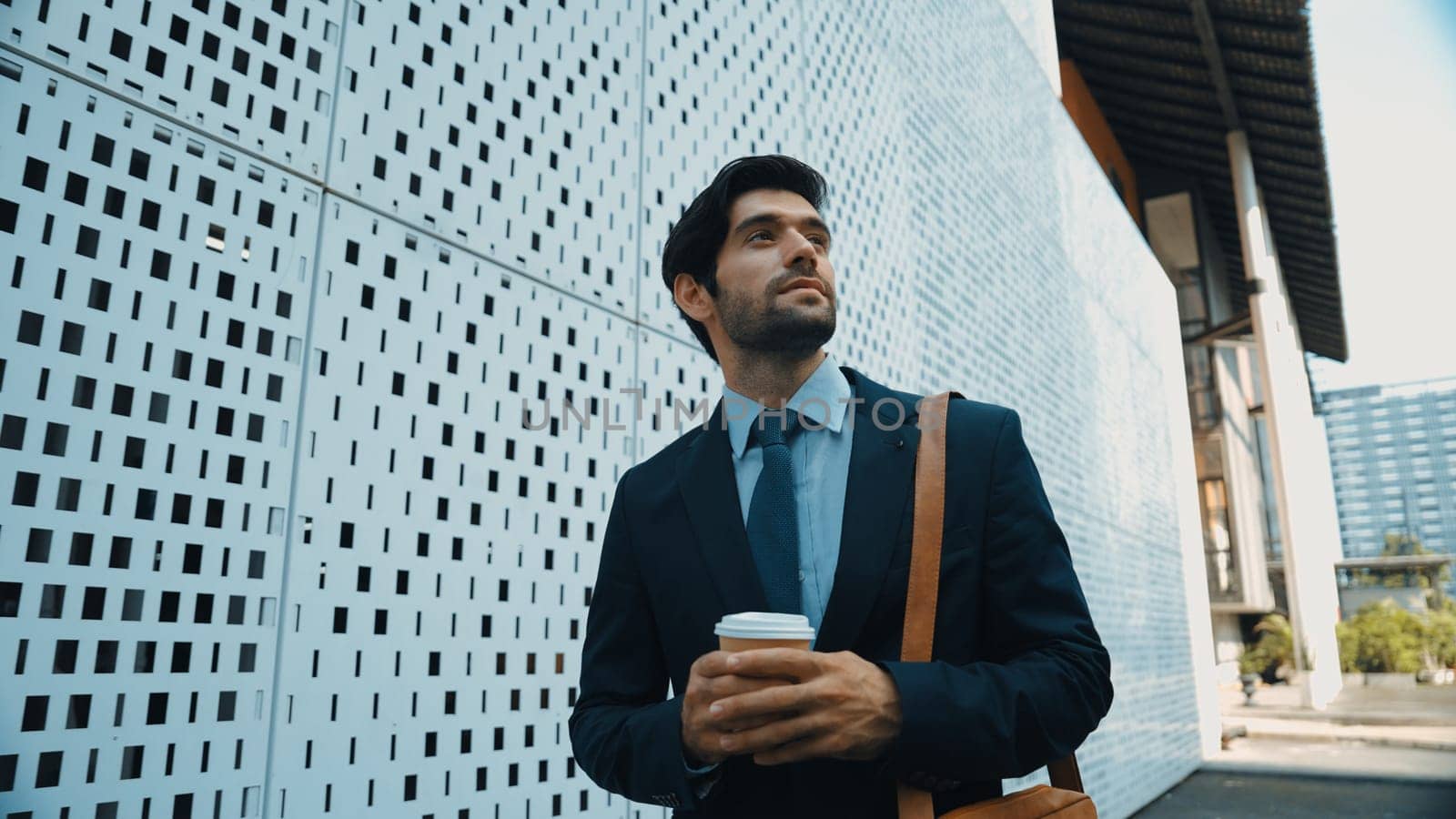 Skilled business man walking in suit outfit while holding coffee cup. Professional manager standing near architectural building. Going to working in morning, seeking for successful job. Exultant.