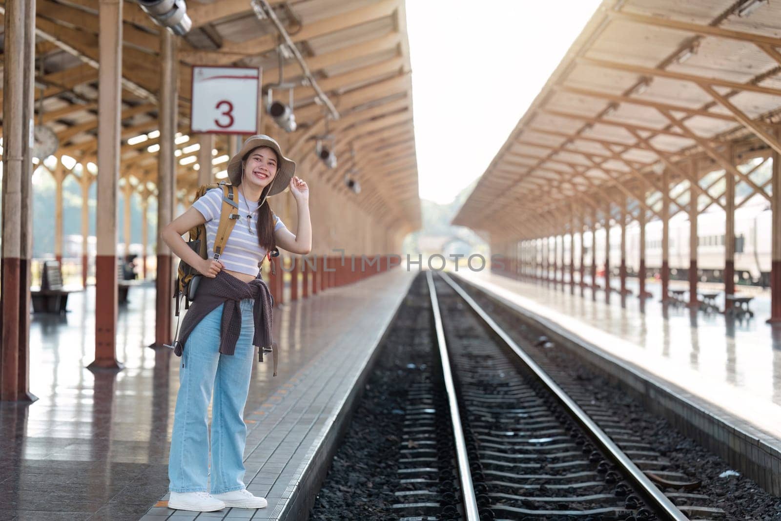 Young Female Traveler at Train Station Platform Ready for Adventure, Wearing Backpack and Hat, Smiling and Looking Excited for Journey, Sunlight Streaming Through Wooden Roof Structure by itchaznong