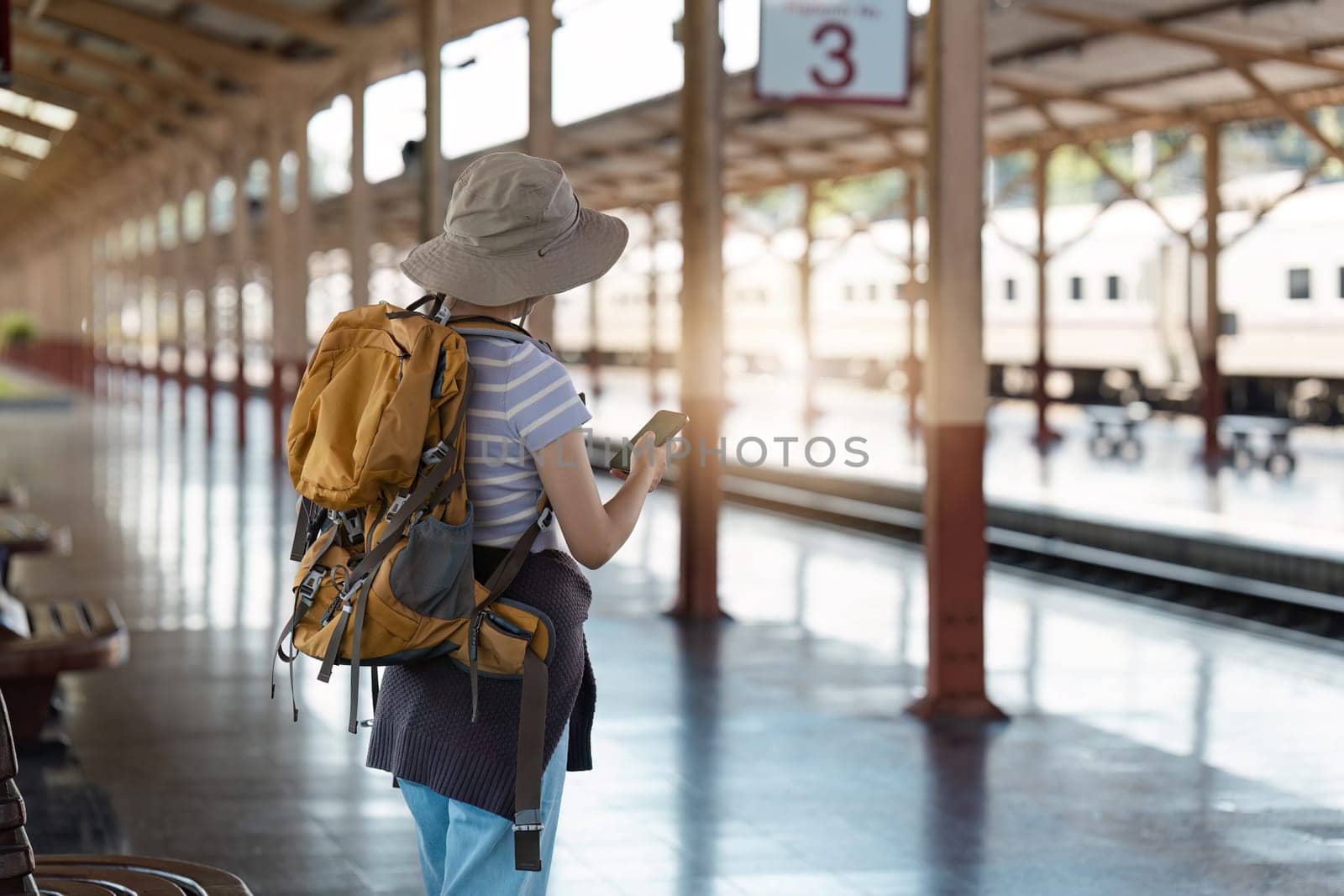 A solo traveler with a backpack and hat stands at a train station platform, checking their phone for travel updates. Perfect for themes of adventure, exploration, and travel.