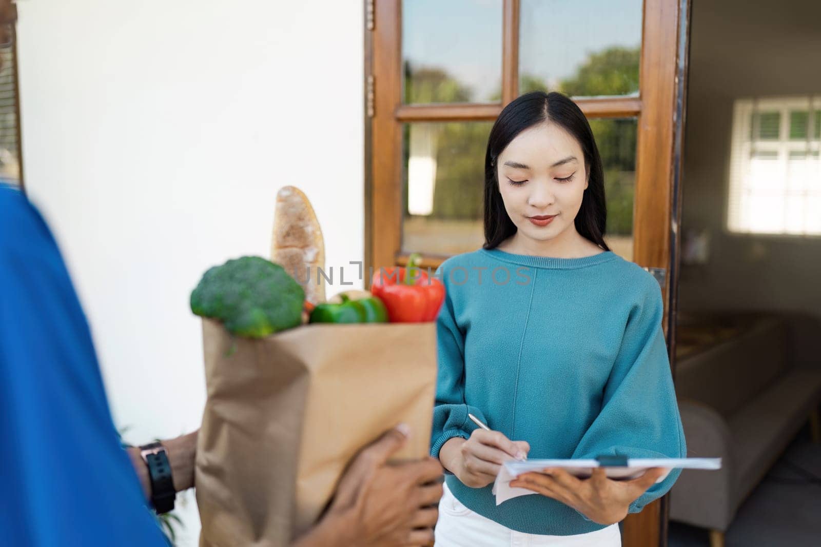 Woman signing for grocery delivery at home, receiving fresh produce from delivery person at her doorstep.