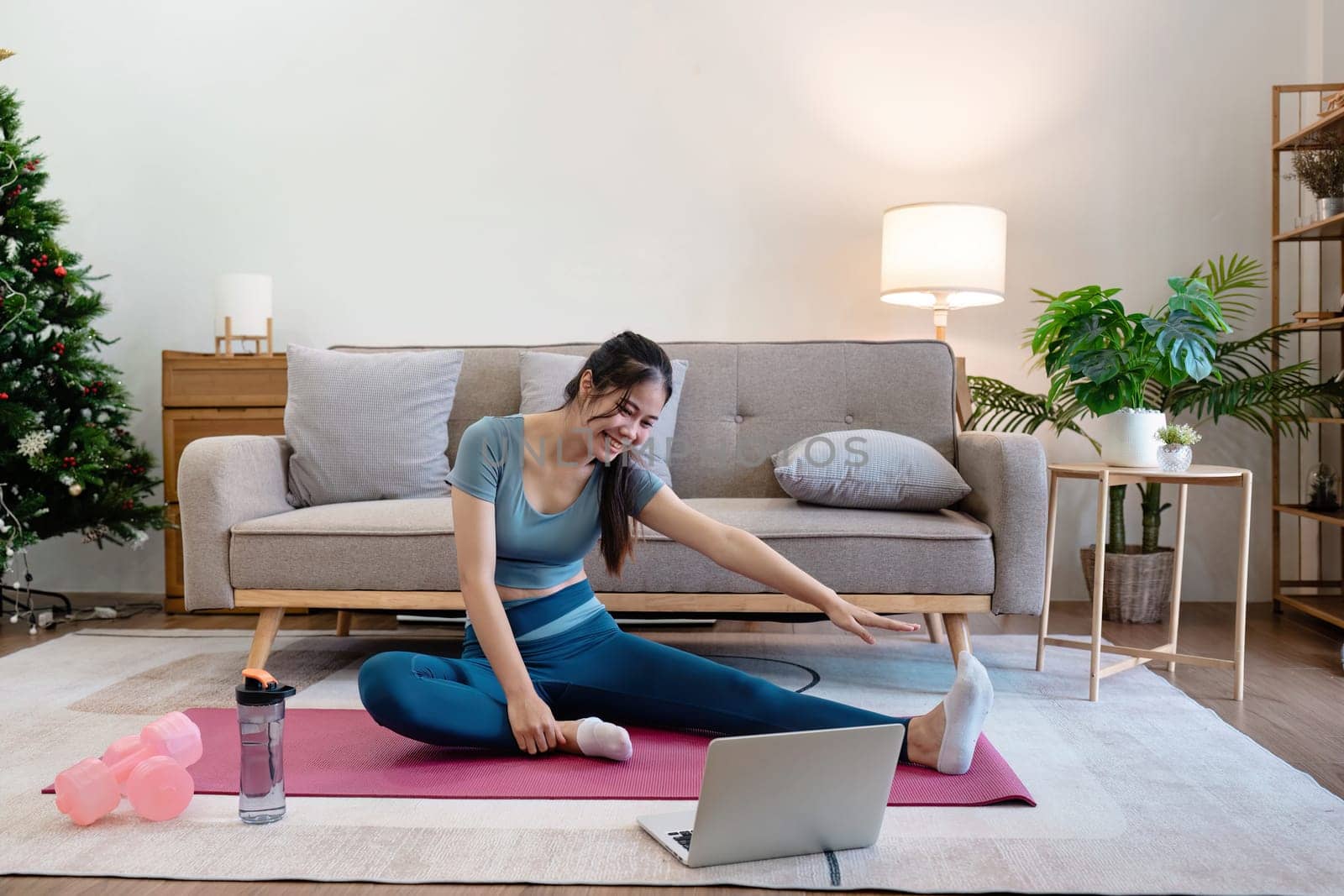 Woman stretching on yoga mat in modern living room with laptop, water bottle, and dumbbells. Cozy and bright interior with sofa, plants, and Christmas tree.