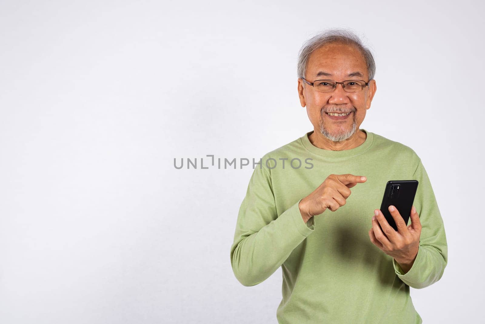 Portrait smiling Asian older man smiling and pointing at his cell phone studio shot isolated on white background. elderly holding mobile phone in hand by Sorapop