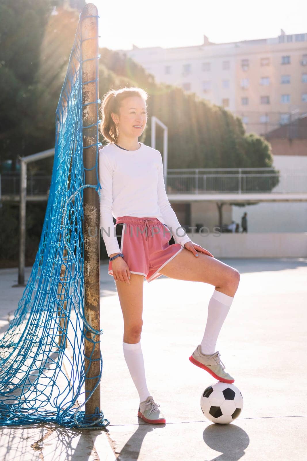 smiling female football player stepping on a soccer ball next to the goal of an urban court, concept of sport and active lifestyle