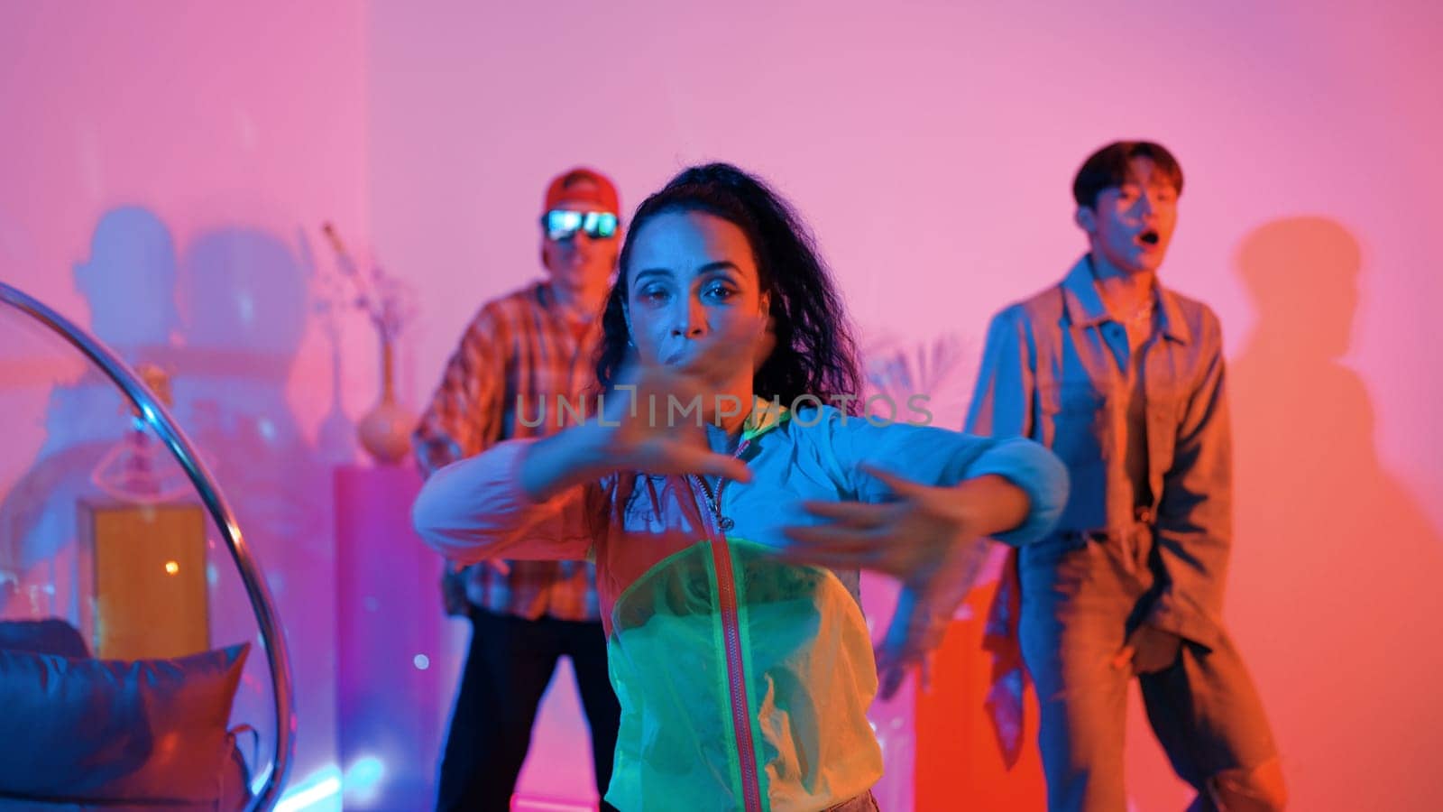 Attractive happy dancer looking at camera while smart woman moving at rhyme with neon light. Professional hispanic performer break dancing while wearing colorful cloth with diverse friend. Regalement.