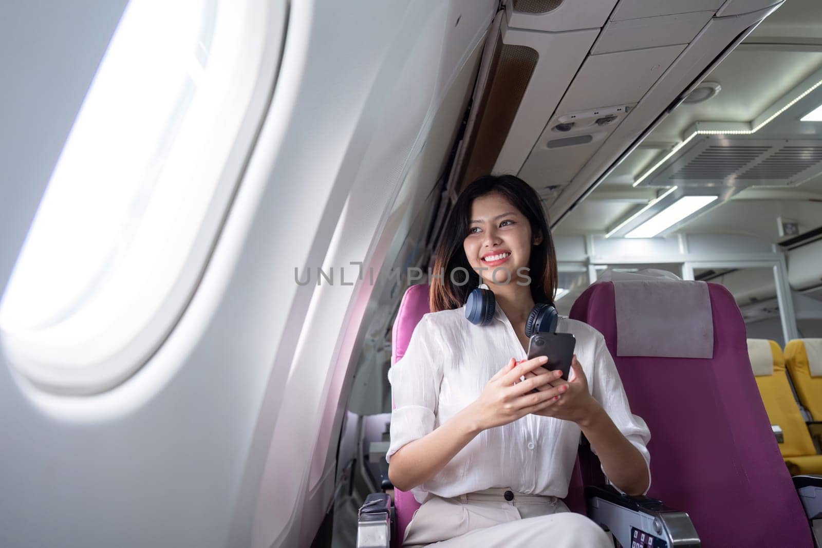 Business Woman Traveling on Airplane, Smiling Professional Female with Smartphone and Headphones, Modern Air Travel, Comfortable Seating, In Flight Experience, Corporate Travel, Business Trip by nateemee