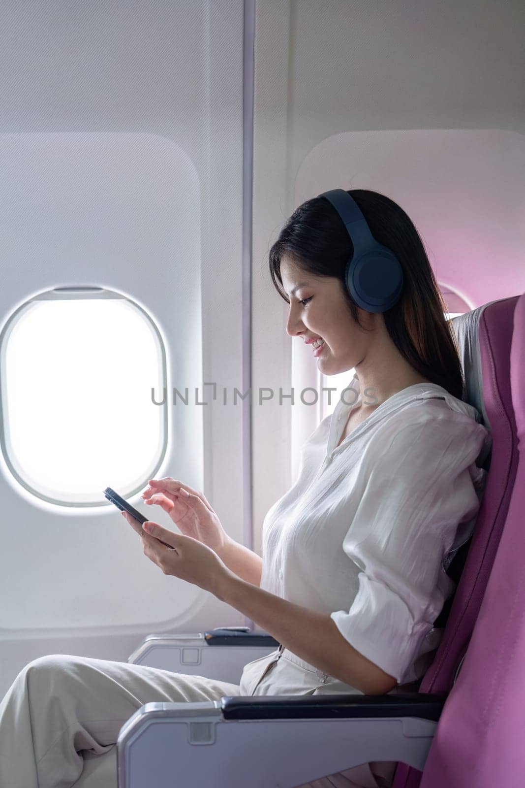 Businesswoman traveling by airplane, using smartphone and wearing headphones, seated by window, enjoying modern air travel.