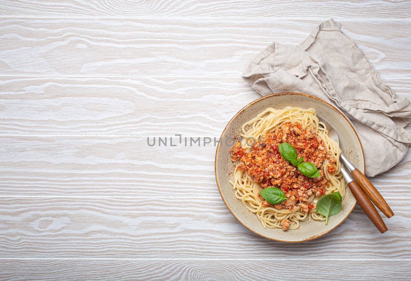 Plate of spaghetti bolognese, topped with fresh basil leaves, sits on a white wooden table with a linen napkin