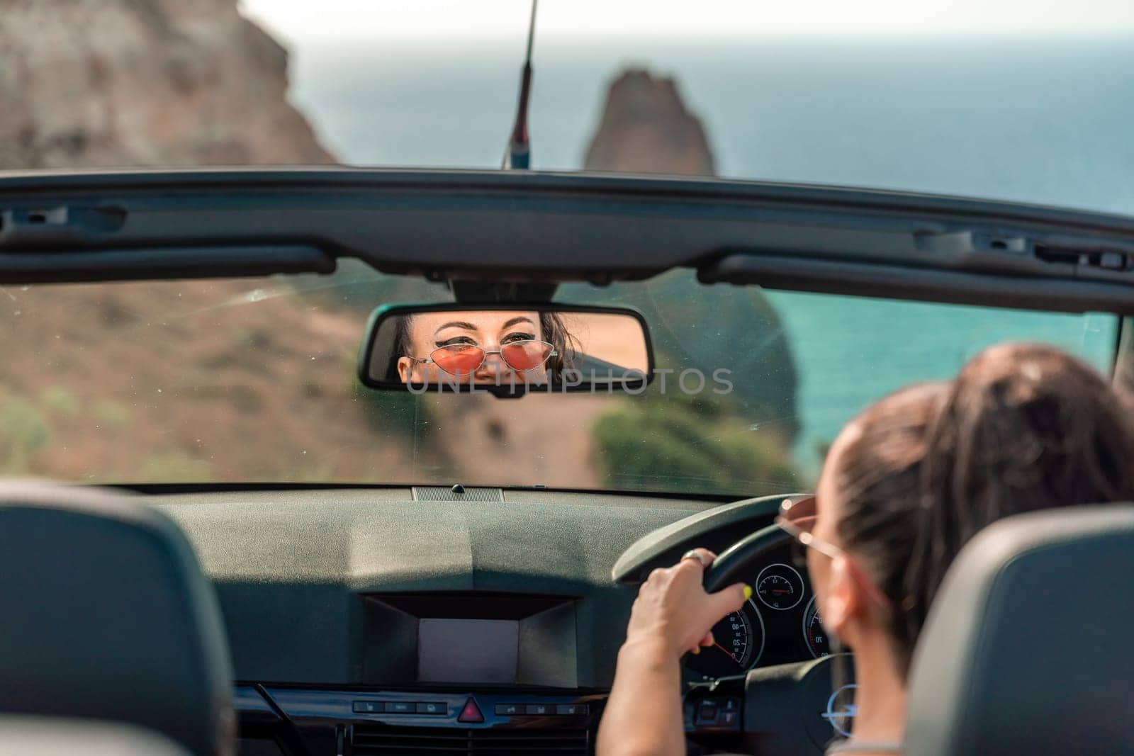 A woman is driving a convertible car with the top down, enjoying the ocean view. The car is red and black, and the woman is wearing a pink shirt