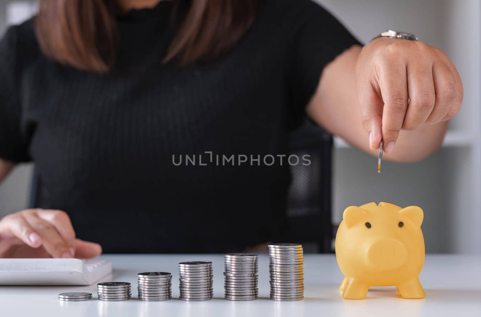 Young Woman Planning to Save Money with Piggy Bank and Calculator, Financial Planning, Budgeting, and Savings Concept, Close-up of Hand Placing Coin in Piggy Bank by wichayada