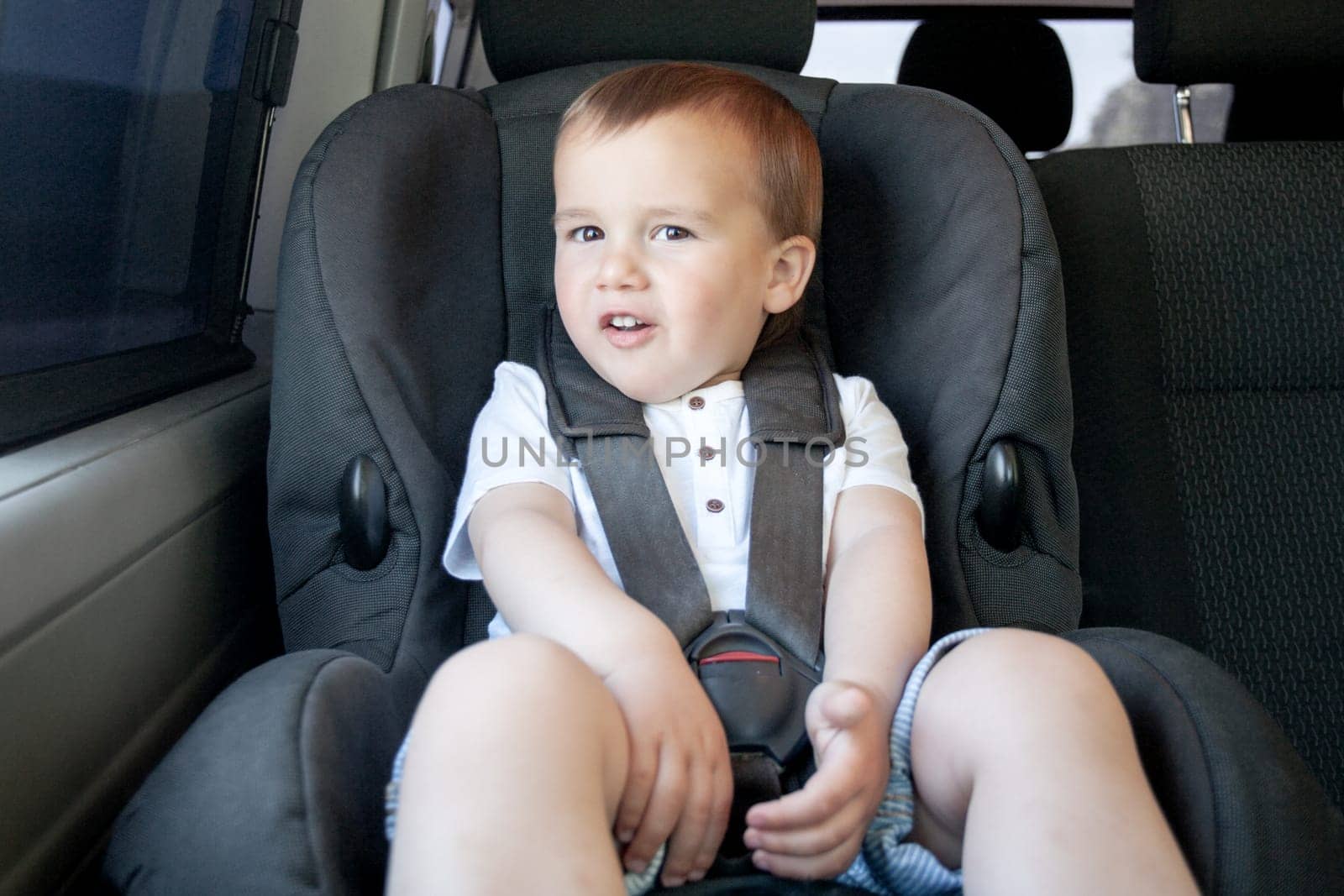 Toddler sitting in child safety seat of car looking at camera. Transportation and safety.  by orafotograf