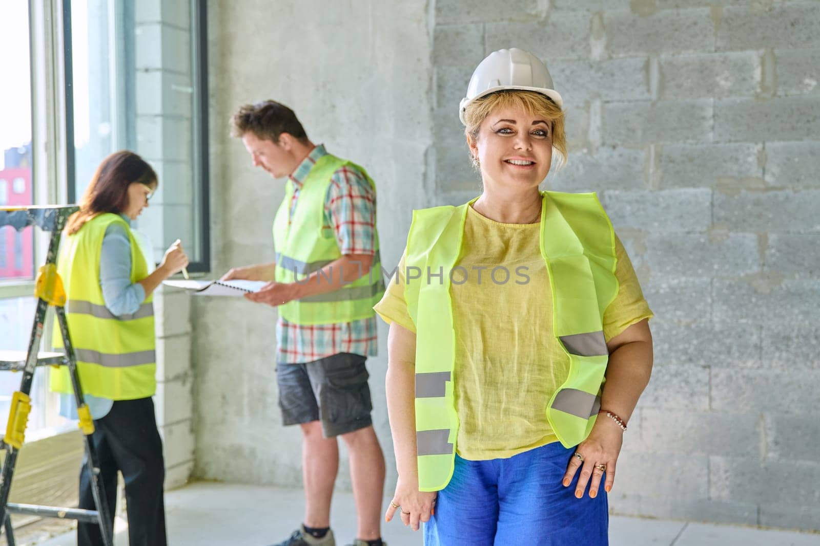 Female designer managing finishing decoration in safety helmet vest looking at camera with team workers in background in cement room of office. Industrial workers construction repair work concept