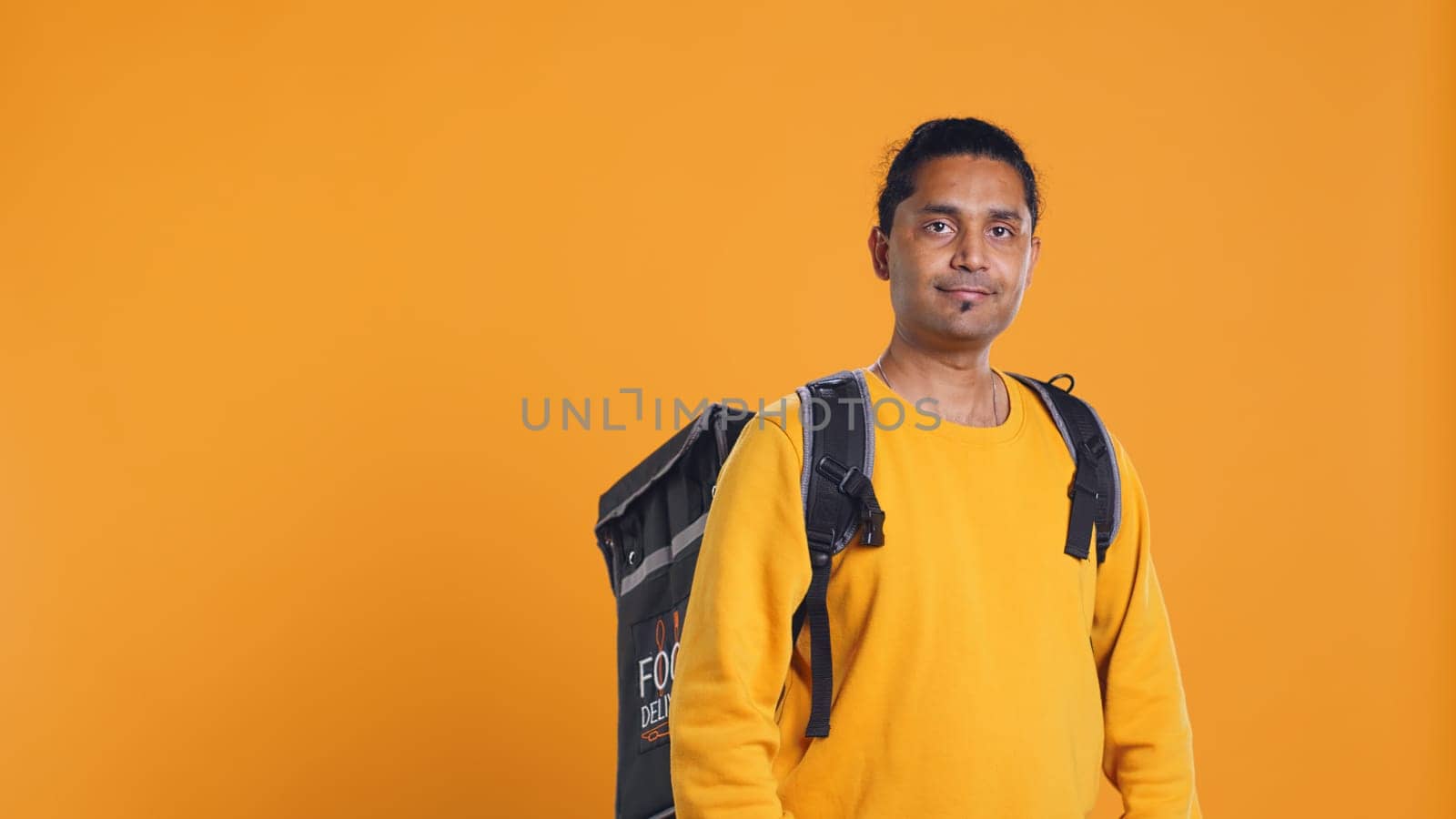 Portrait of happy smiling indian bike rider holding thermal backpack, isolated over studio background. Friendly courier prepared to deliver takeaway food to customer, camera B