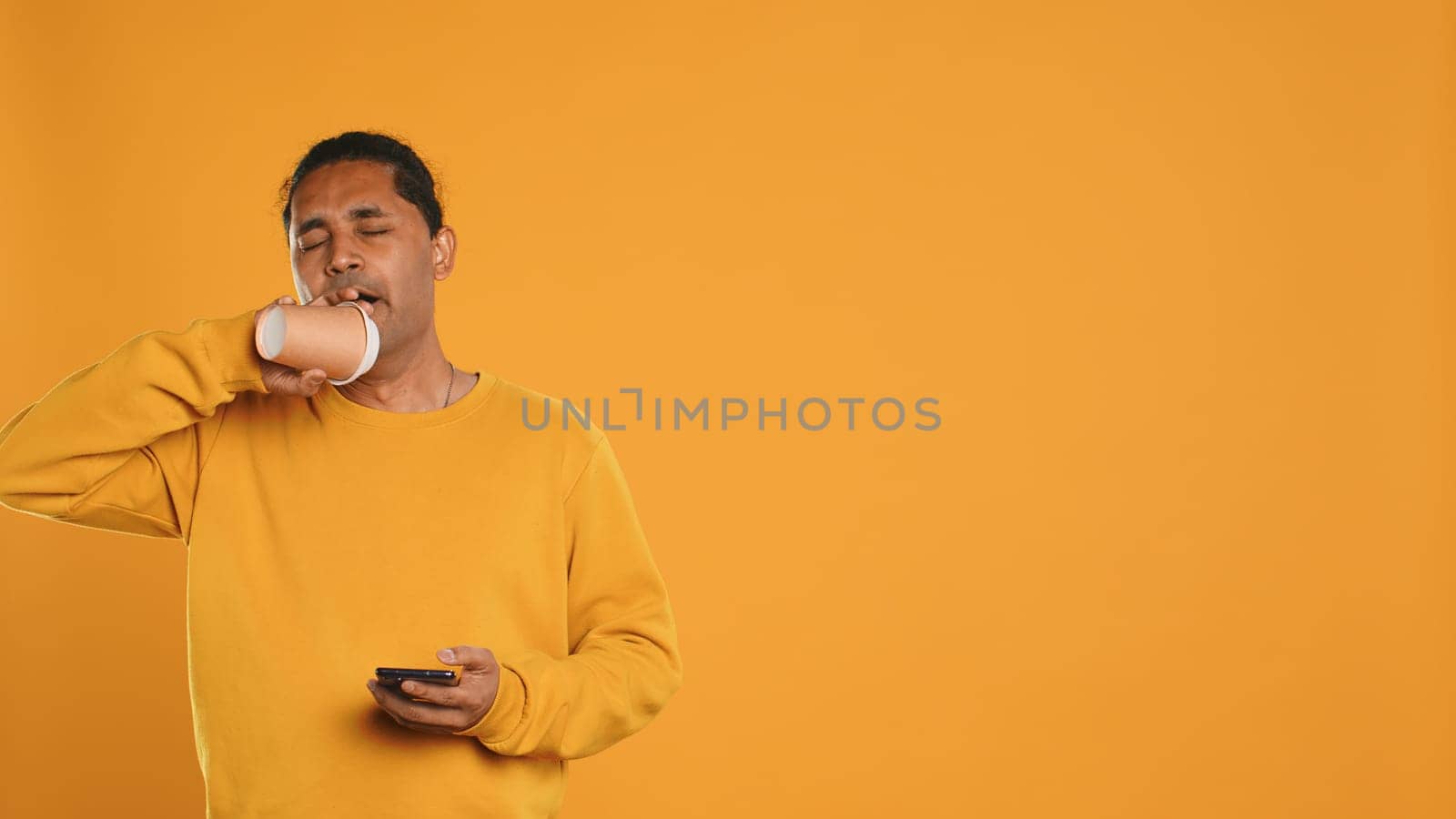 Indian man enjoying fresh coffee from disposable paper cup early in the morning to wake up and be energized. Person drinking hot beverage from recycled takeaway cup, studio background, camera A