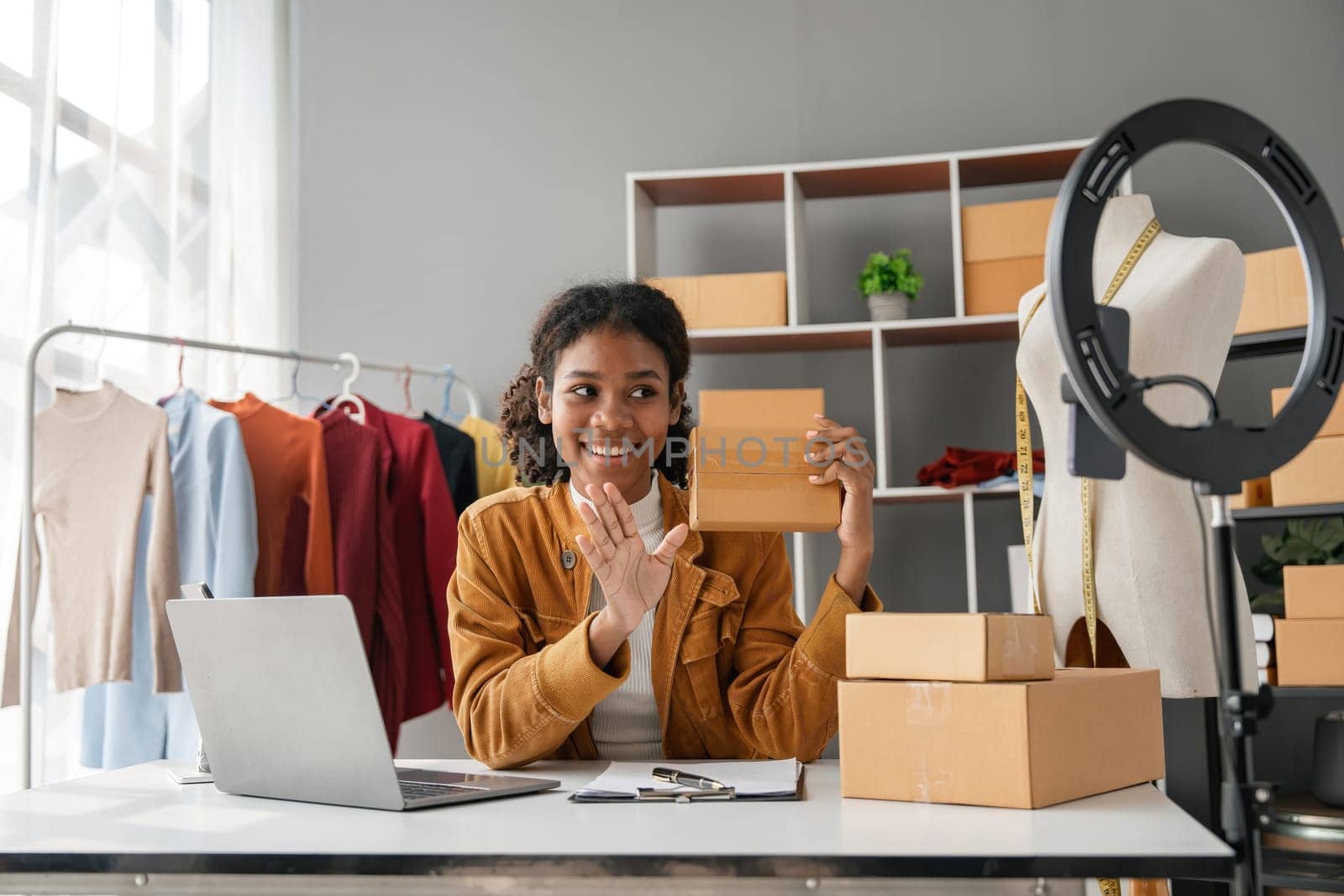 Young female entrepreneur managing her online store, packing orders, and working from her home office. Modern e-commerce business setup with clothing and accessories.