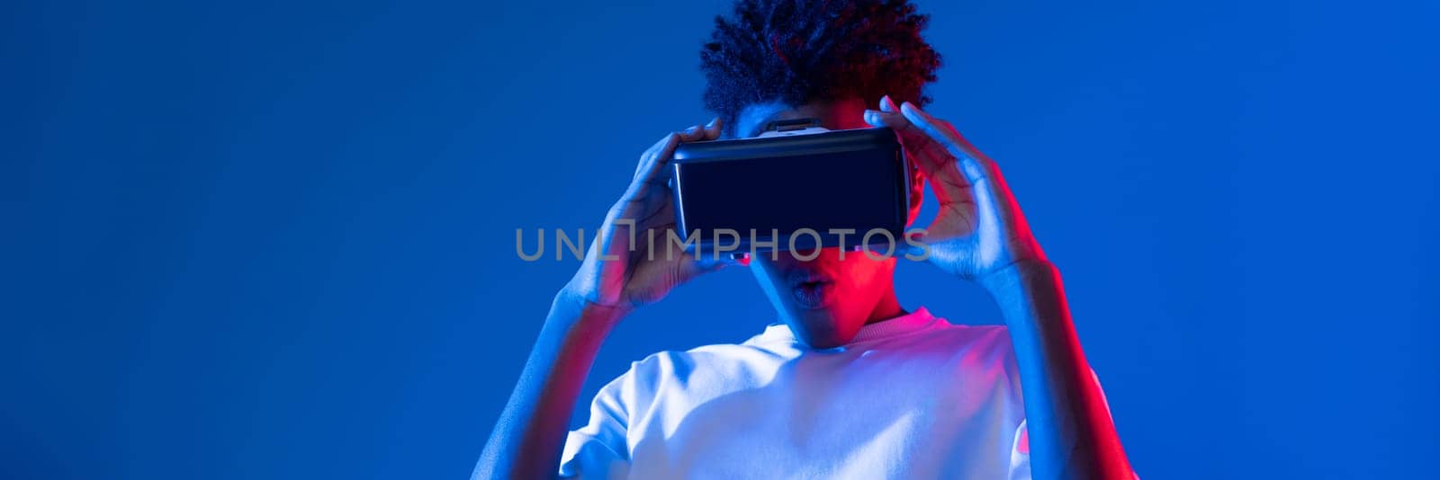 Surprised young African American looking through VR holding interesting object hologram on pink blue neon wall at metaverse world connecting digital futuristic technology virtual reality. Contrivance.