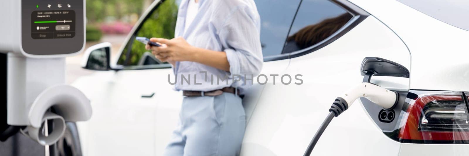 Young woman using smartphone online banking application to pay for electric car battery charging from EV charging station during vacation road trip at national park or summer forest. Panorama Exalt