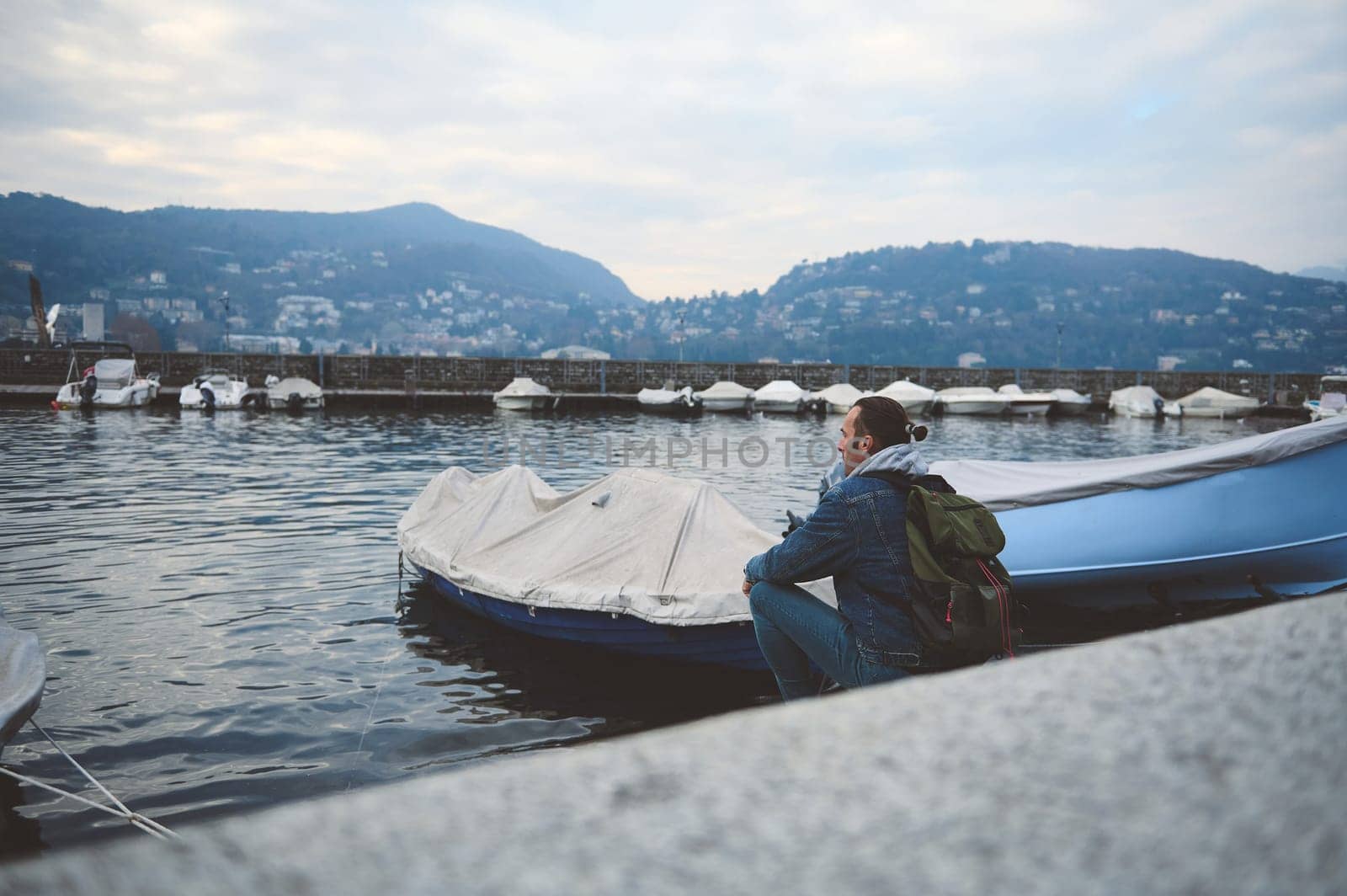 Man sitting by a docked boat at the harbor with mountains in the background by artgf