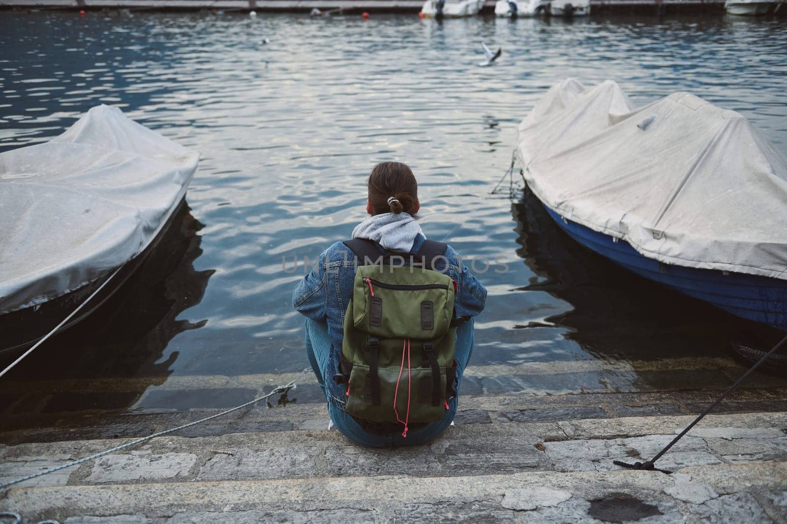 A person with a backpack sitting on stone steps at a dock between two covered boats overlooking a calm water body, reflecting peace and solitude.