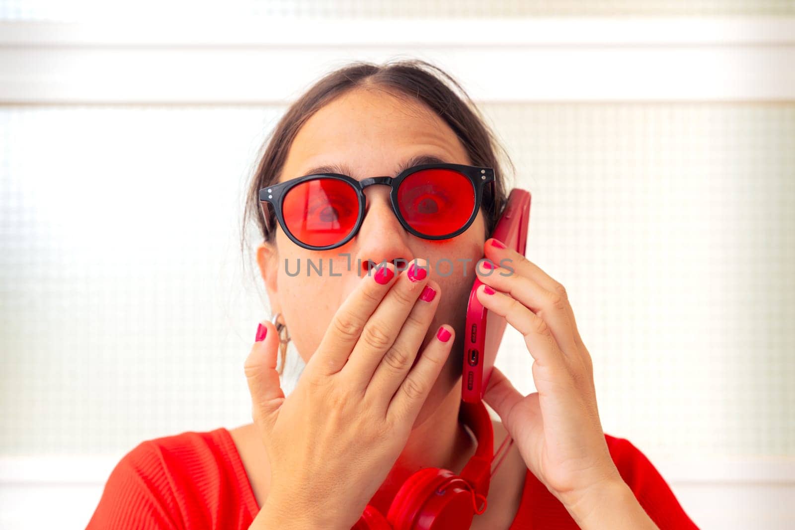 Young woman wearing sunglasses and red dress covering her mouth with her hand when talking on the phone with a friend and laughing