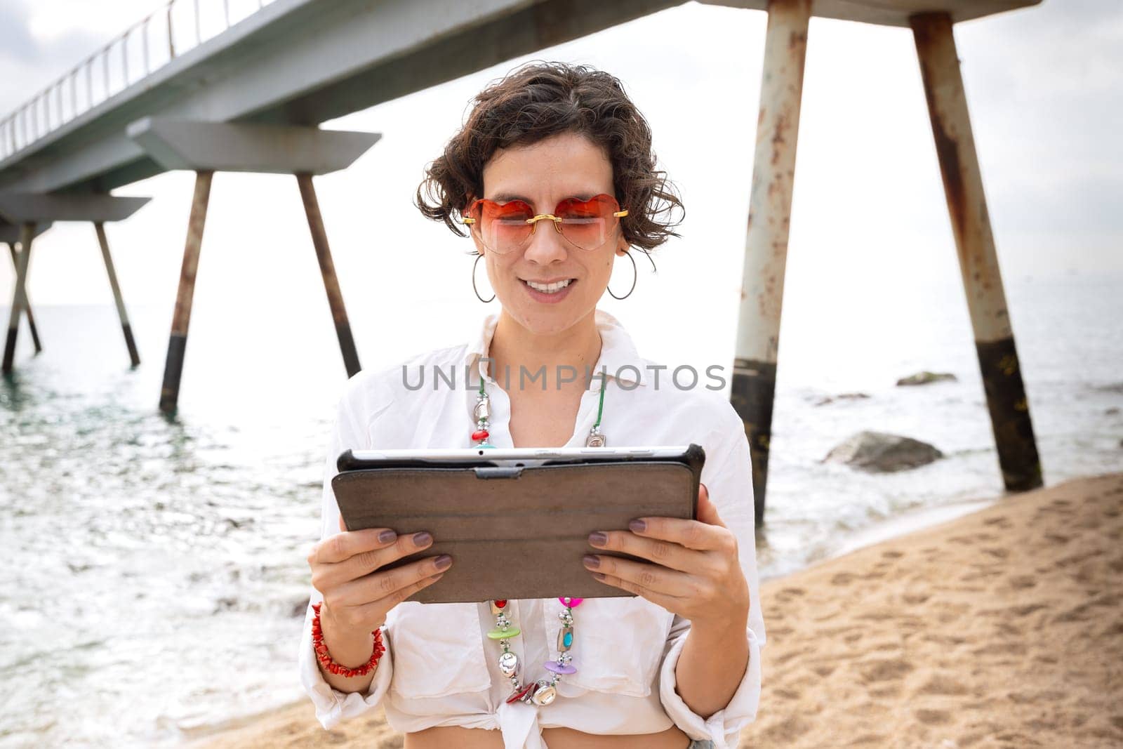 Latin woman wearing sunglasses on vacation by the sea laughing using a social media app with her tablet.