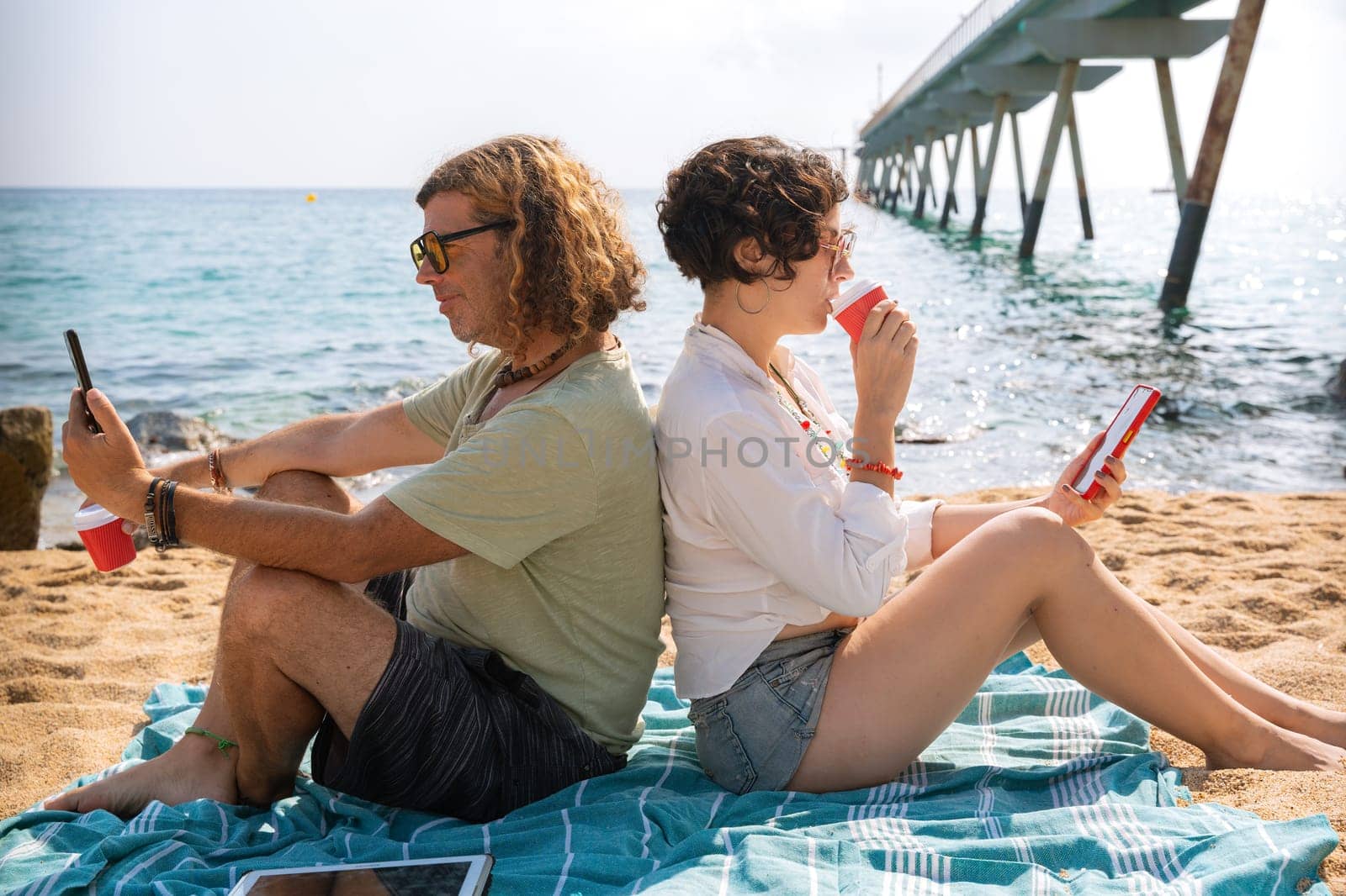 Middle-aged man and woman sitting on the beach with surprised face, browsing telephones apps. Concept:Holidays y Technology