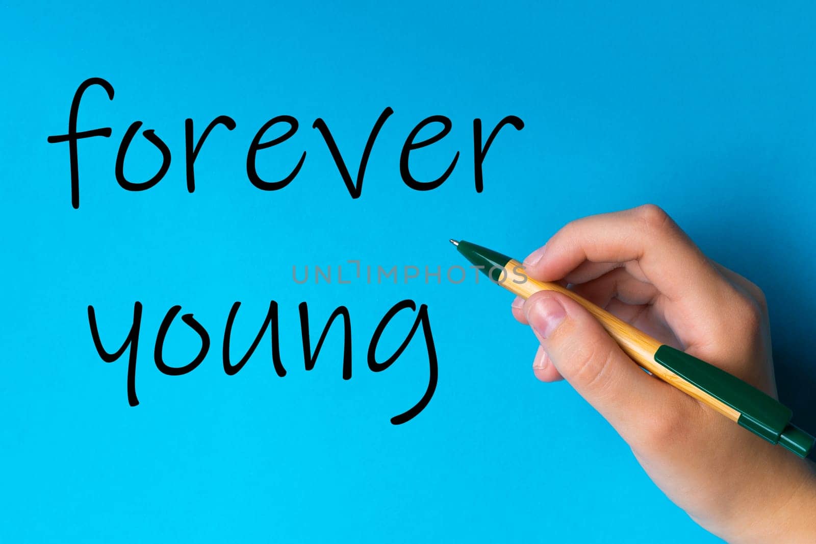 Children hand with pen write on a blue white background. Writing hand. Word Forever young