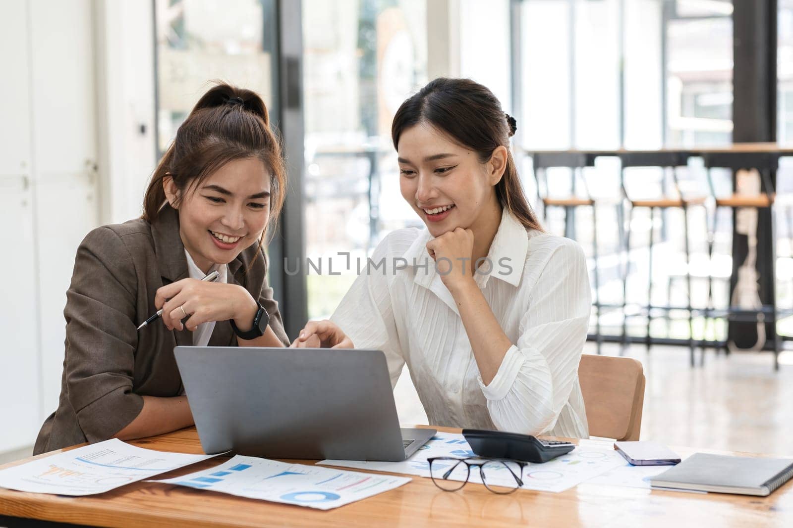 Young Businesswoman Collaborating with Colleague in Modern Office Setting, Discussing Ideas and Analyzing Data on Laptop, Professional Teamwork, and Corporate Collaboration by wichayada