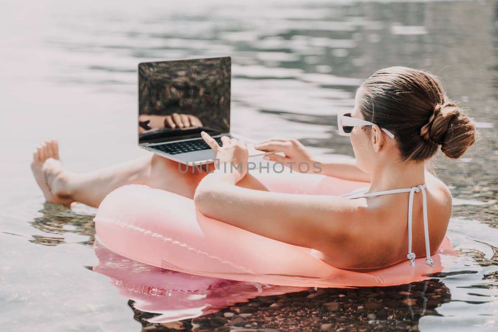 A woman is floating on a pink inflatable raft in the ocean while using a laptop. Concept of relaxation and leisure, as the woman enjoys her time in the water while working on her laptop